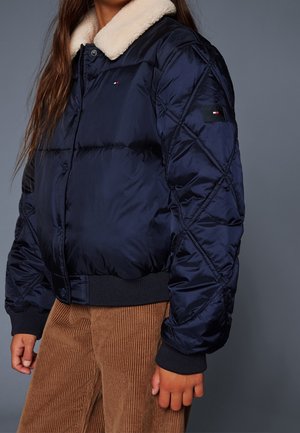 Žena v námořnicky modré prošívané bundě s bílým plyšovým límcem a hnědých manšestrových kalhotách na jednobarevném šedém pozadí.