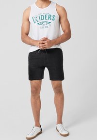 Chemise blanche sans manches avec texte graphique turquoise, associée à un short noir et des baskets blanches aux semelles marron. Tenue simple et décontractée.
