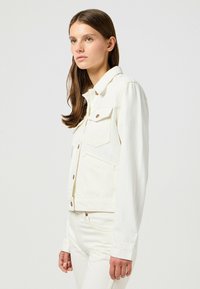Vit denimjacka med långa ärmar, knäppning med knappar och bröstfickor, med en ren design och detaljer i kopparfärg.