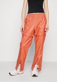 Orange faux leather pants with an elastic waistband, featuring snap button details and side slits. Paired with white sneakers.