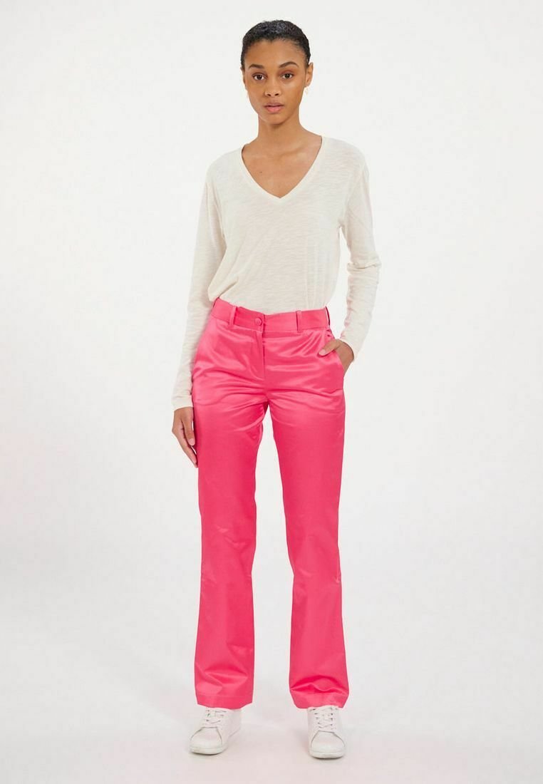 Pantalons taille haute rose vif avec une texture lisse, associés à un haut beige clair à col en V à manches longues. Le mannequin porte des baskets blanches.