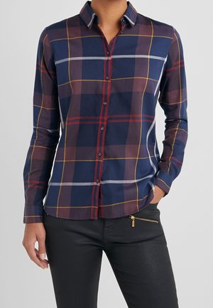 Kobieta ubrana w koszulę w kratę w granatowym, czerwonym i żółtym kolorze oraz czarne spodnie, z jedną ręką w kieszeni, na tle jasnoszarej gładkiej ściany.