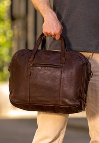 Brown leather briefcase with zippered pockets, dual handles, and a smooth texture. Features stitching and a subtle logo design.