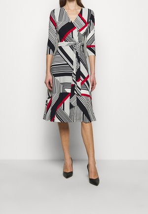 Vrouw dragen een wikkeljurk tot op de knie met zwart-witte en rode geometrische strepen, driekwartmouwen, en zwarte puntige hakken.