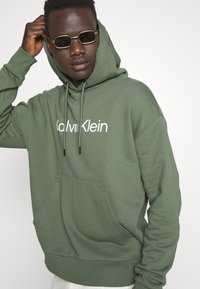 Felpa verde con cappuccio, tasca frontale e coulisse, con il logo bianco "Calvin Klein" sul petto. Texture in misto cotone liscia.