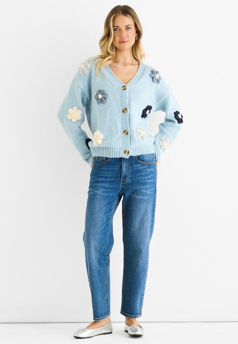 Light blue knitted cardigan with floral appliqués in cream, navy, and gray, featuring a V-neck, button front, and ribbed cuffs. Paired with blue jeans.