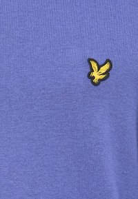 Violetter gestrickter Pullover mit strukturiertem Finish und einem kleinen schwarzen und goldenen Adlerlogo auf der linken Seite.
