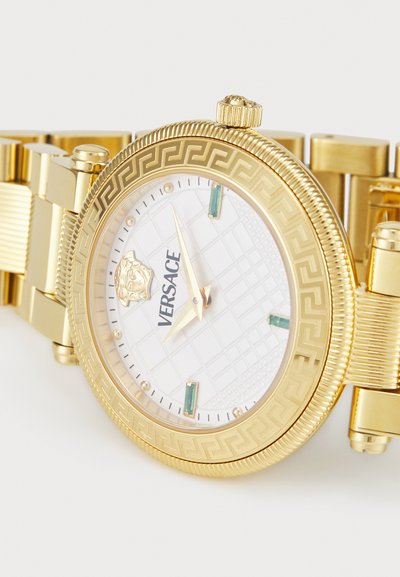 Auksinės spalvos „Versace“ laikrodžio su balta raštuota ciferblatu, žaliais akcentais ir tekstūruotu rėmeliu. turi metalinę apyrankę ir logotipų dekoracijas.