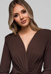 Maglietta marrone a maniche lunghe con scollatura a V profonda e dettaglio frontale attorcigliato. Tessuto morbido; il modello ha capelli mossi e orecchini, con trucco neutro.