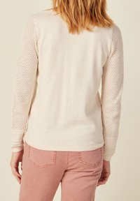 Ljusbeige tröja med långa ärmar som har ett texturerat stickat mönster på ärmarna. Kombinerad med rosa byxor. Mjuk tyg, avslappnad design.