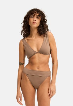 Kobieta z kręconymi ciemnymi włosami, ubrana w taupe bikini z górą w stylu kopertowym i majtkami z wysokim stanem na jednolitym jasnym tle.