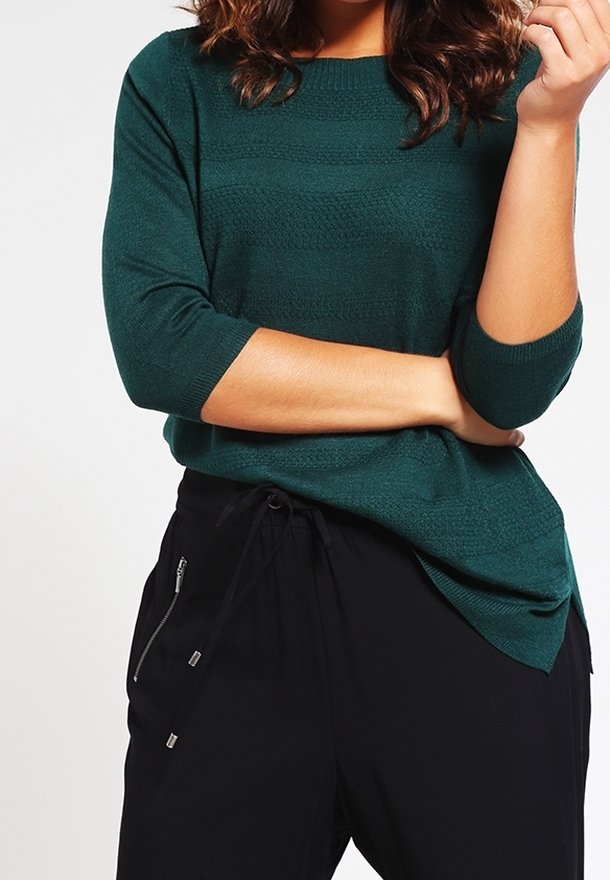 Femme portant un pull texturé bleu sarcelle à manches trois-quarts et un pantalon noir à cordon avec poche zippée, bras croisés.