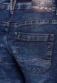 Granatowe dżinsy z jasnym kontrastowym przeszyciem i widoczną tylnią kieszenią. Posiadają metkę z napisem "DENIM" i "NIGDY MNIE NIE OPUŚĆ."