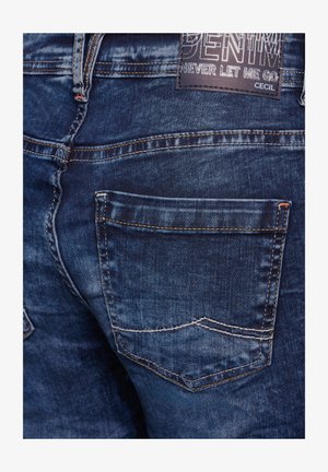 Donkerblauwe denim jeans met lichte kontraststiksels en een zichtbare achterzak. Bevat een stoffen label met de tekst "DENIM" en "NEVER LET ME GO."