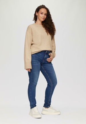 Beżowy sweter z prążkowanymi mankietami, w połączeniu z ciemnoniebieskimi jeansami slim fit i białymi trampkami, stojący na neutralnym tle.