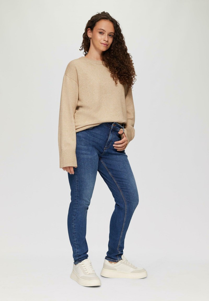 Beige trui met geribbelde manchetten, gecombineerd met donkere blauw slim-fit jeans en witte sneakers, staand tegen een neutrale achtergrond.
