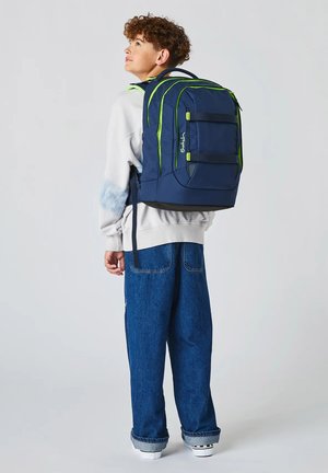 Teenager mit lockigem Haar, der einen weißen Pullover und blaue Jeans trägt, trägt einen großen dunkelblauen Rucksack mit grünen Akzenten.