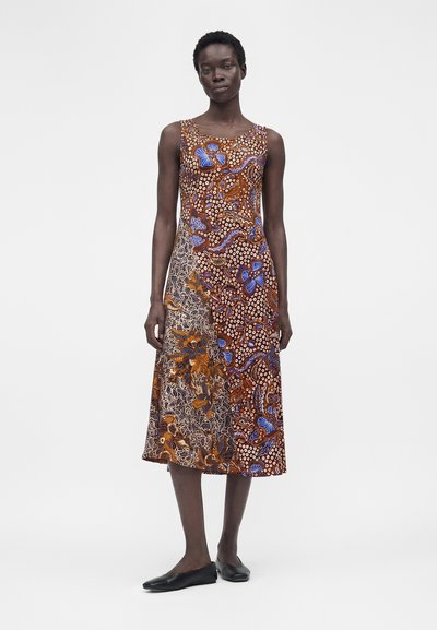 Γυναίκα στέκεται φορώντας αμάνικο φόρεμα μέχρι τον αστράγαλο με ανάμεικτα καφέ, μπλε και πορτοκαλί λουλουδάτα και αφηρημένα σχέδια, συνδυασμένο με μαύρες μπαλαρίνες.