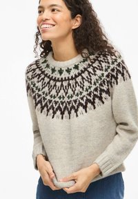 Kobieta uśmiechająca się, ubrana w beżowy sweter z dzianiny z ciemnobrązowymi i zielonymi geometrycznymi wzorami wokół dekoltu, trzymająca rękami dół swetra.