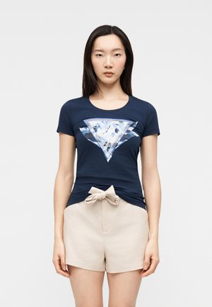 Donna con capelli lunghi e scuri che indossa una maglietta blu navy con un motivo a triangolo lucido e pantaloncini beige con fiocco frontale.