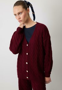 Sieviete, kas valkā bordo krāsas adītu džemperi ar pogām un atbilstošām biksēm, ar tumši zilu kreklu un rakstainu matu šalli.