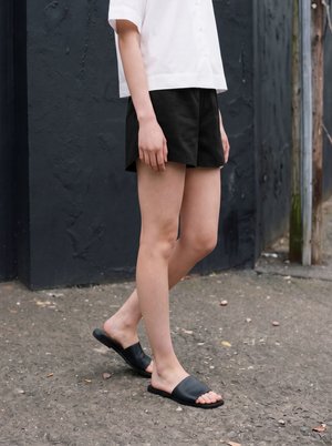 Person iført sorte slidssandaler, sorte shorts og en hvid kortærmet skjorte står på ru asfalt nær en mørk væg.