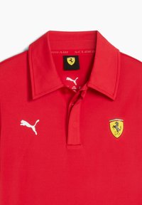 Un polo rouge avec un logo Puma blanc et un emblème Ferrari représentant un cheval cabré et un blason jaune. Conception classique du col.