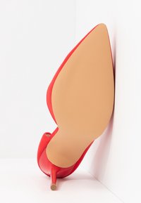 Chaussure à talon haut rouge vue de dessous avec une semelle pointue et un talon aiguille sur un fond blanc en coin.
