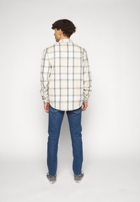 Homme portant une chemise à carreaux beige et noire à manches longues, un jean bleu et des baskets grises, debout devant un fond blanc.