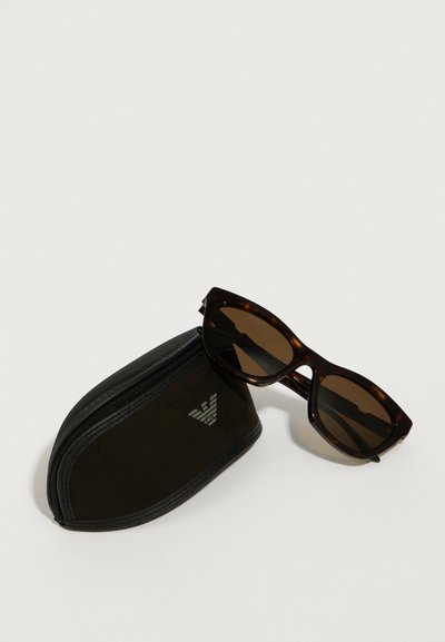 Ochelari de soare tortoiseshell maro cu lentile închise, așezați lângă o husă neagră cu fermoar, având un logo argintiu pe o parte.