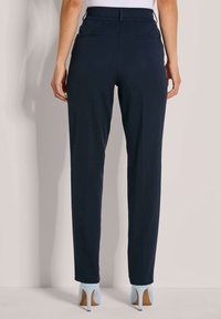 Vrouw in marineblauwe slim-fit broek met elastische tailleband en lichtblauwe hoge hakken, staand tegen een neutrale achtergrond.