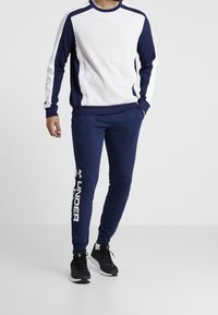 Felpa grigia e blu con polsini a coste, abbinata a pantaloni jogger blu con logo bianco "Under Armour". Sneakers nere con suola in gomma.