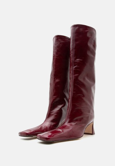 Ψηλές μπορντό δερμάτινες μπότες με χαμηλό τετράγωνο τακούνι και τετράγωνη μύτη, σε όρθια θέση σε απλό λευκό φόντο.