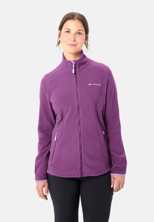 Frau trägt eine lila Fleecejacke mit Reißverschluss und zwei Reißverschlusstaschen, steht vor einem schlichten hellen Hintergrund.