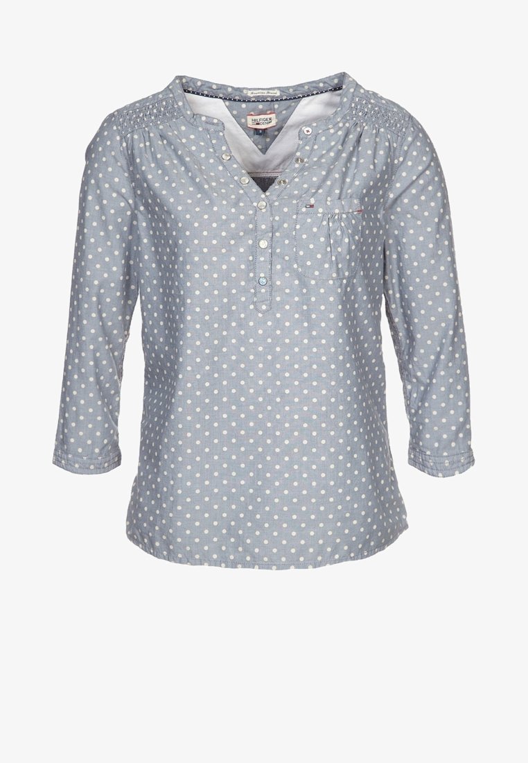 Blouse bleu clair à manches longues avec pois blancs, col en V boutonné, poche poitrine et détails froncés aux épaules.