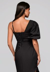 Schwarzes Kleid mit einem mit Pailletten verzierten Oberteil, asymmetrischem Ein-Schulter-Design und strukturiertem Puffärmel. Glatte Textur mit einem hohen Schlitz bis zur Oberschenkel.
