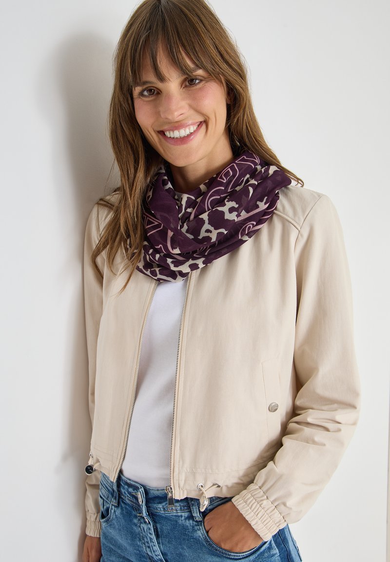 Jacket bomber beige con zip frontale, polsini elastici e orlo con cordino. Abbinata a una sciarpa modellata viola e bianca.
