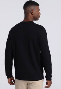 Black long-sleeve shirt with a textured fabric, rounded neckline, and a relaxed fit, worn with beige pants. Features long sleeves and a straight hem.
