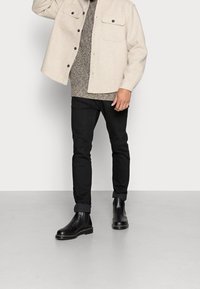 Giacca camicia in misto lana beige con due tasche sul petto, maglione grigio a trama, pantaloni slim neri e stivali Chelsea neri in pelle con suola spessa.