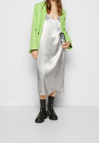 Robe en satin argenté, blazer oversize vert et bottines noires à chunky. Le blazer a une finition texturée, tandis que la robe a une surface lisse.