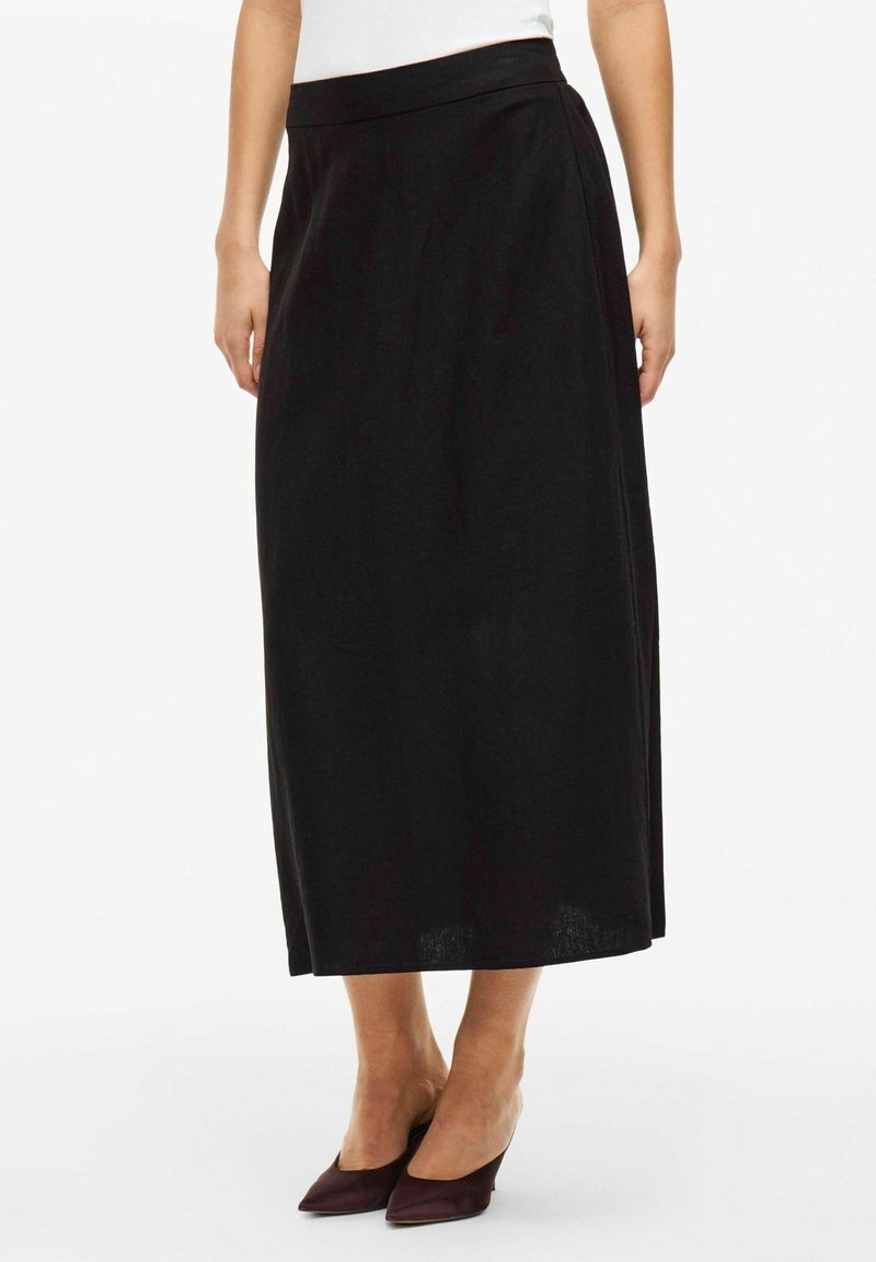 Femme portant une jupe noire, taille haute, longueur cheville avec une coupe droite, associée à des escarpins pointus foncés.