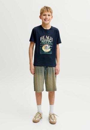 Niño rubio sonriendo, lleva una camiseta azul marino con la palabra "BEACH" y un diseño tropical, pantalones cortos verdes descoloridos hasta la rodilla, calcetines blancos y zapatillas beige.