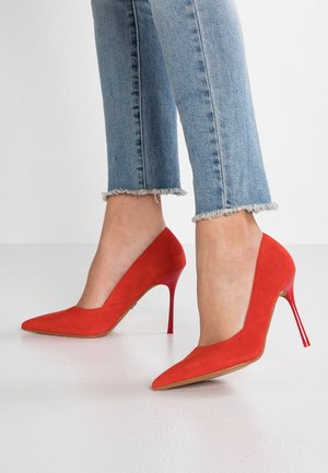 Rode suède hoge hakken met een spitse neus, voorzien van een dunne rode stilettohak en een slank ontwerp. Gekozen in combinatie met blauwe jeans met een rafelige zoom.