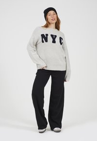 Kobieta w szarej bluzie z NYC, czarnych szerokich spodniach, białych butach i czapce beanie, stoi z jedną ręką w kieszeni na białym tle.