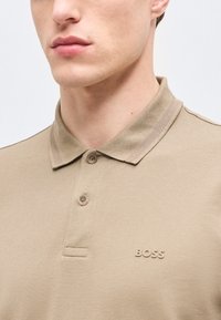 Άνδρας που φοράει μπεζ πόλο μπλουζάκι με δύο κουμπιά και λογότυπο "BOSS" στο αριστερό στήθος, μπροστά σε λευκό απλό φόντο.