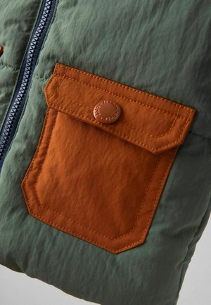 Detailný záber na zelenú látkovú bundu s oranžovou zasúvacou vreckom a viditeľným čierno-bielym zipsom na ľavej strane.