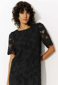 Robe en dentelle noire avec broderie florale, manches courtes, col rond et tissu texturé. Présente des bords à volants.