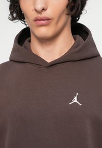 Joven con piel clara que lleva una sudadera con capucha marrón oscuro con un pequeño logo Jumpman blanco en el pecho.