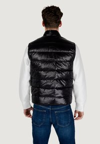 Chaleco acolchado negro con acabado brillante, con secciones acolchadas horizontales. Llevarlo sobre una camisa de manga larga blanca, combinado con jeans azules.
