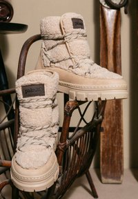 Stivali beige con tomaia in tessuto peloso e dettagli in suede, dotati di suola in gomma robusta e lacci decorativi. Patch con logo sul lato.